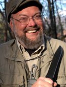 Picture of a bearded man with glasses, wearing a hat and a heavy coat wielding a knife outdoors.