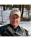 Picture of a clean-shaven man wearing a white ATF hat in front of a snowy forest